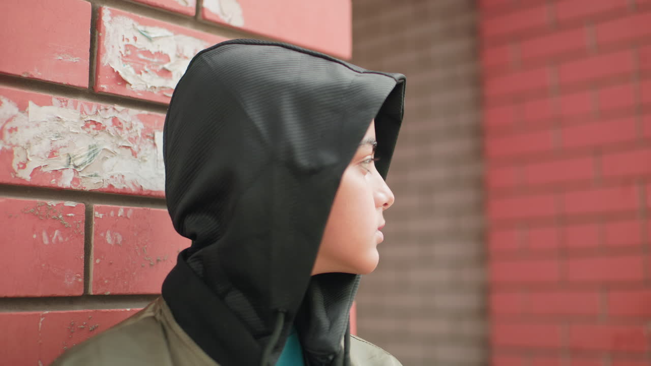 Little boy in hooded jacket standing against brick wall gradually lifting head up with eyes closed thoughtfully, calm expression captured in close up with urban background