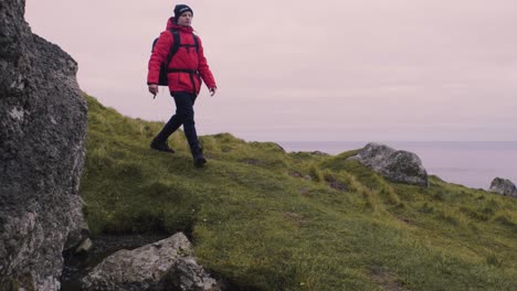 man in red jacket and with a backpack on hikes through the green hills and big rocks on the beautiful mountain trek on the faroe islands