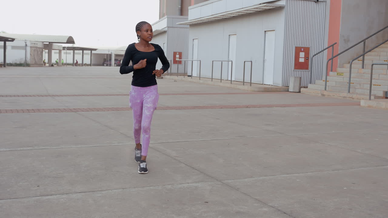 Donna che corre attraverso una piazza di cemento vuota, indossando un top nero e leggings viola. Mantiene un passo di allenamento costante vicino a edifici industriali all'alba. Appassionata di fitness concentrata che si riscalda, con le sue scarpe da ginnastica che battono sull'asfalto.