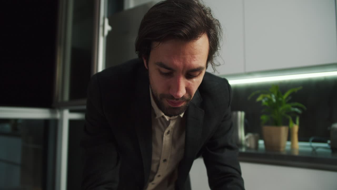 Serious brunette man with stubble wipes sweat from his forehead in a black jacket and beige shirt working in front of his laptop and then rejoicing at his success moving away from his desk showing his beige shorts in a modern apartment in the evening