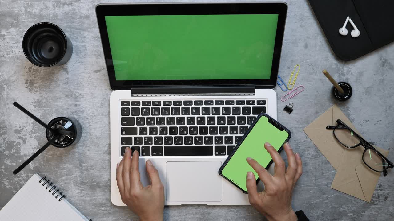 Businesswoman using smartphone with green screen at office table. Modern office work desk with laptop computer and office accessories. Female fingers zooming, tapping on smartphone green screen.