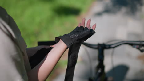 vista trasera de primer plano de un individuo asegurando el guante de motociclista en la mano derecha al lado de la bicicleta, con enfoque en los dedos ajustando la correa, el fondo muestra vegetación borrosa y camino pavimentado