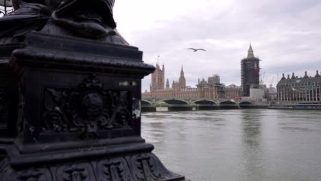 View-of-Westminster-parliament-and-Big-Ben-under-renovation-work-and-construction,-from-the-other-side-of-the-river-Thames,-birds-flying-in-slow-motion,-tourism-in-London-on-a-cloudy-day