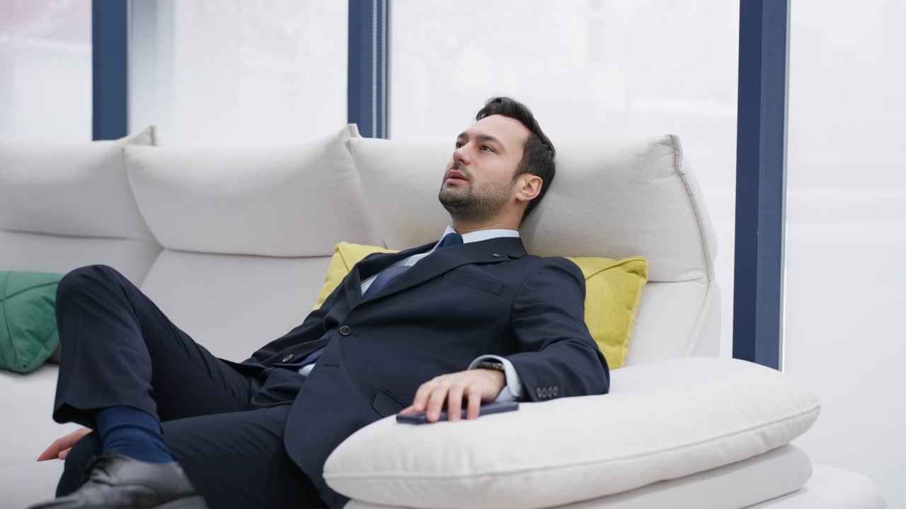A man in a formal suit reclines relaxing casually on a white sofa with colorful cushions in the background, set against large windows. Businessman relaxing on couch at home smiling