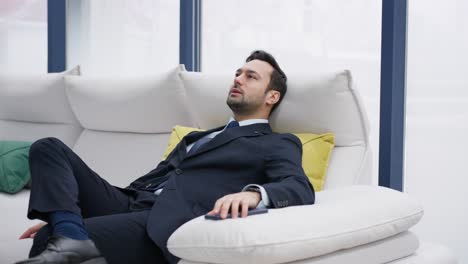 a man in a formal suit reclines relaxing casually on a white sofa with colorful cushions in the background, set against large windows. businessman relaxing on couch at home smiling