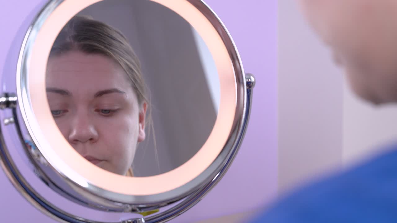 Jovem mulher caucasiana atraente com grandes olhos azuis aplica sombra ao redor da linha dos cílios, usando cores neutras, olhando para seu reflexo em espelho redondo. Maquiagem diária moderna. Em ambiente interno.