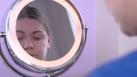 joven mujer caucásica atractiva con grandes ojos azules aplica la sombra de los ojos alrededor de la línea de las pestañas, usando colores neutrales, mirando su reflejo en el espejo redondo. maquillaje diario moderno. en el interior.