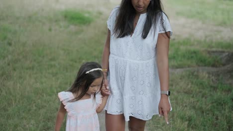 a pregnant woman and her young daughter, both dressed in white, walk hand-in-hand through a grassy park. the scene captures a serene moment of family bonding in nature, surrounded by trees.