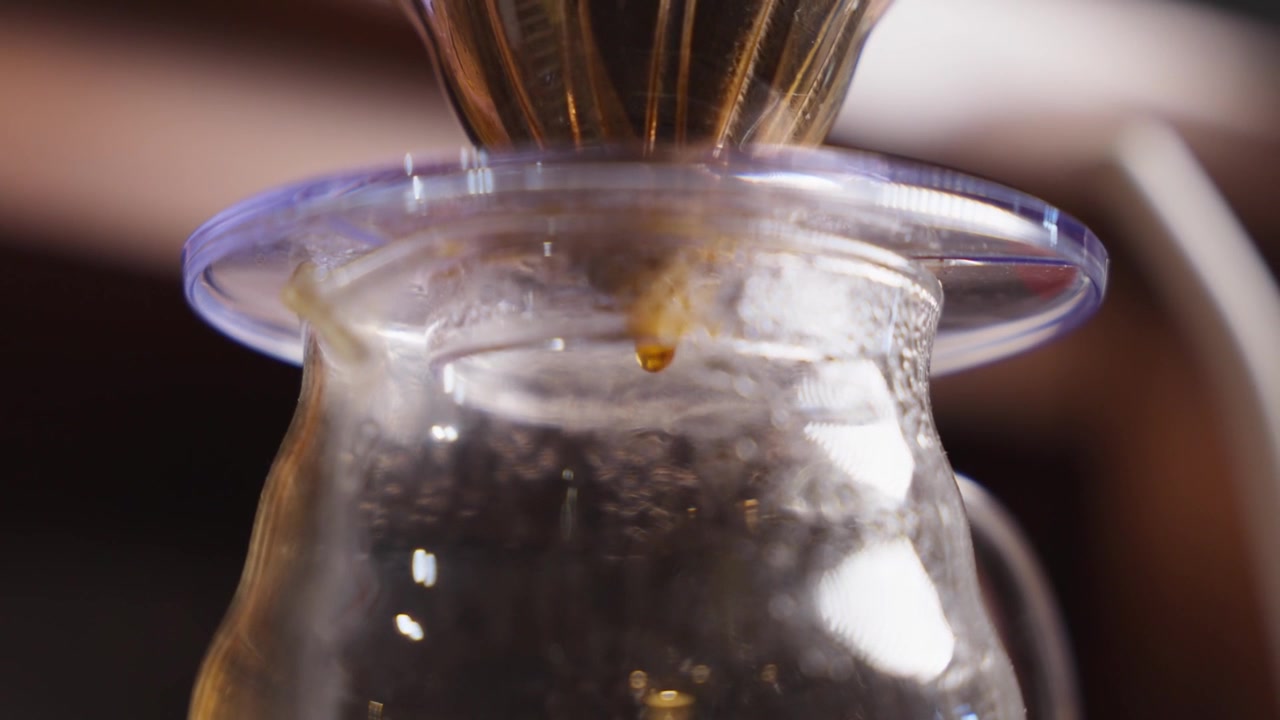 Close-up of pour-over coffee dripping through a paper filter into a glass carafe, showing rich brew color, slow coffee preparation and warm cafe atmosphere.