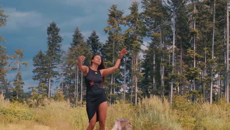 Front-view-of-single-elated-girl-wearing-short-dark-blue-dress-dances-twirling-outdoors-in-nature-pine-tree-forest-background-at-dusk,-blue-sky,-handheld-full-shot