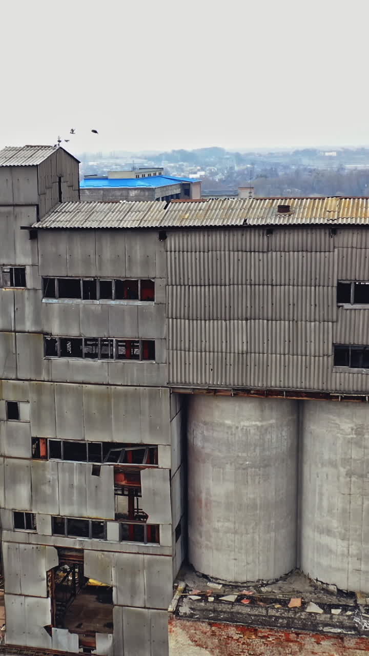 A huge state-owned factory with gray walls and broken glasses in the windows on the background of city buildings. Industrial zone. Aerial view. Vertical video