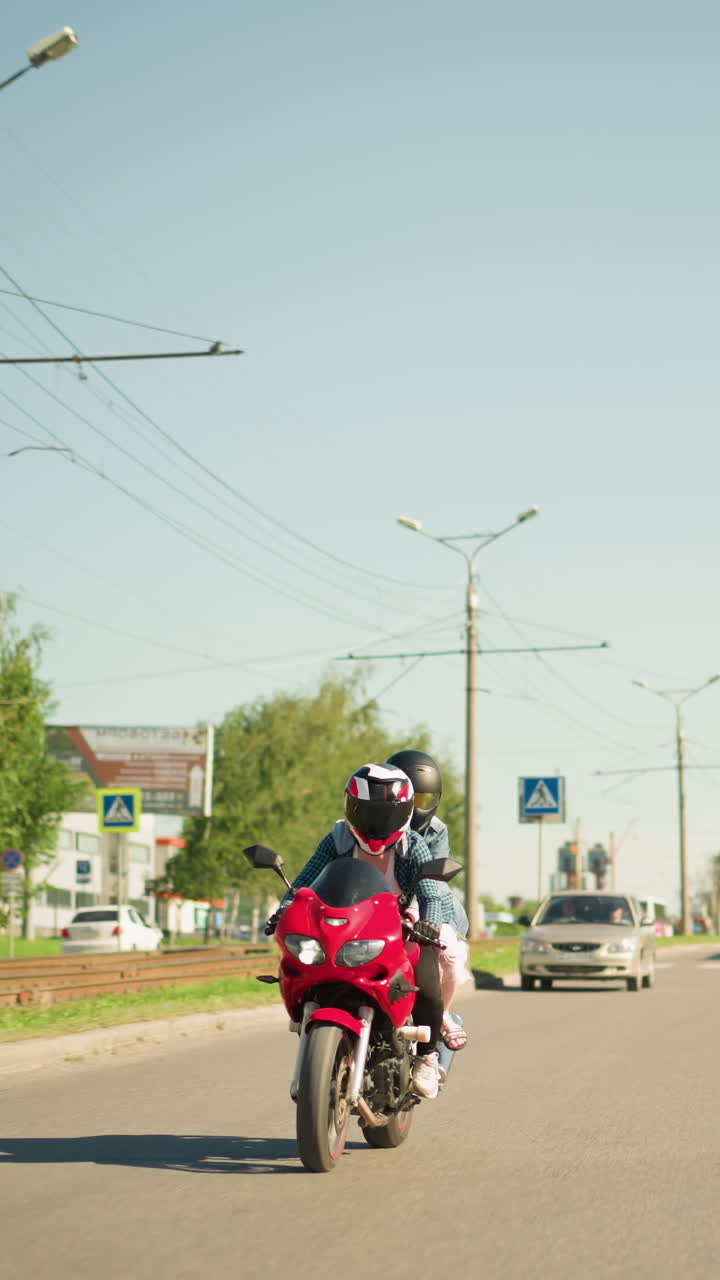 두 친구, 둘 다 헬을 입고, 도시 도로를 따라 빨간색 전력 자전거를 함께 타고, 차와 버스가 뒤에서 따라오고, 도로 건너편에 표지판과 건물이 보입니다.