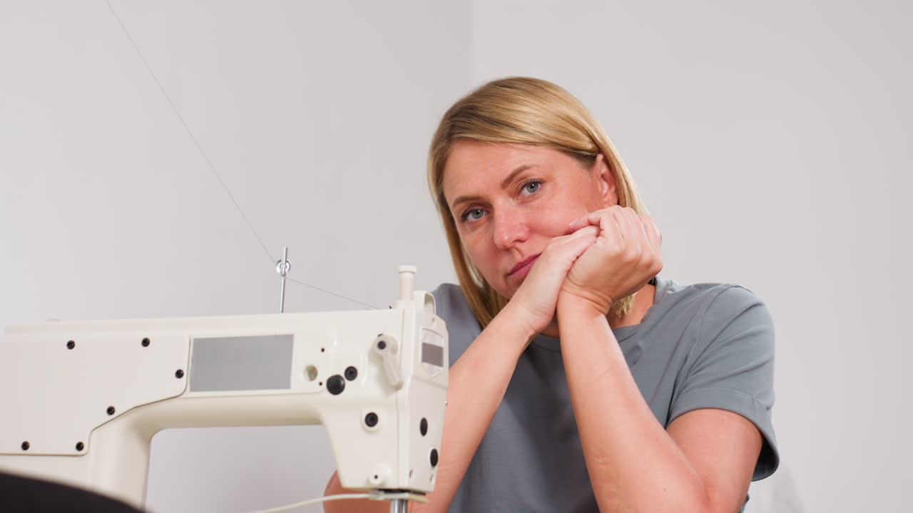 Creative individual resting hands on mouth at table, leaning toward machine, deeply thinking about design choices, concept development, in garment creation, displaying thoughtful approach