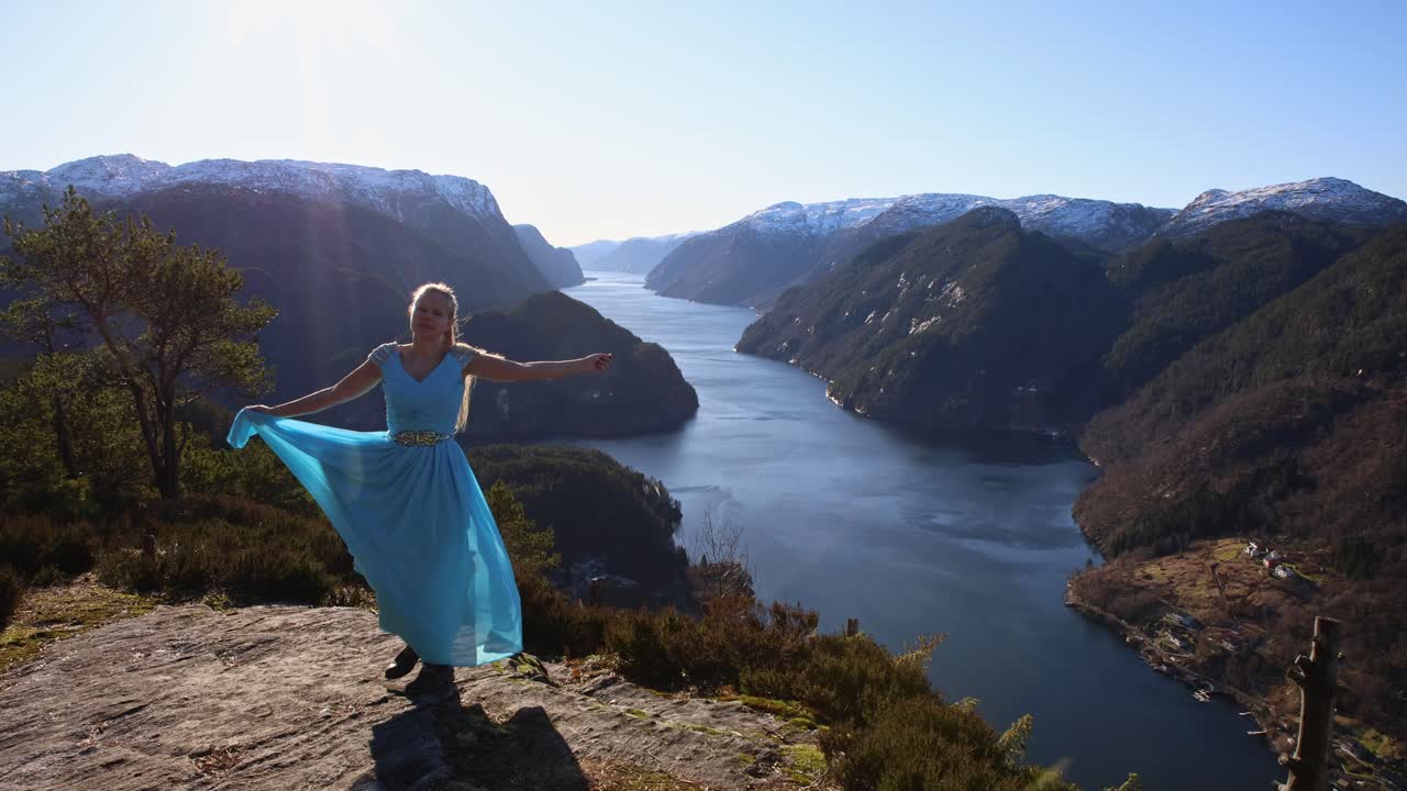 Young woman in blue dress walks to edge of bluff overlooking Norwegian fjord, spins and twirls in both directions, walks off camera.