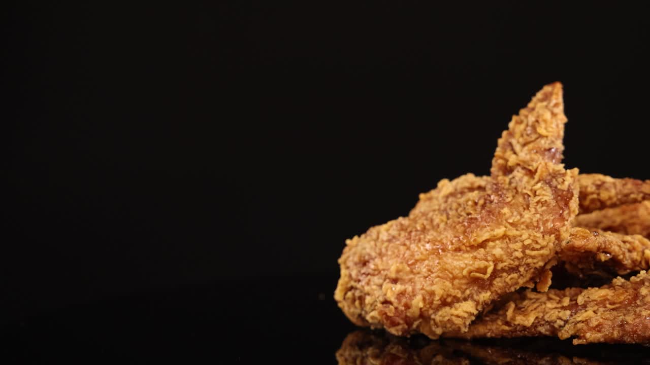 Golden, crispy fried chicken wings rotate smoothly on a glossy black surface with dramatic studio lighting, highlighting texture and detail in a seamless loop