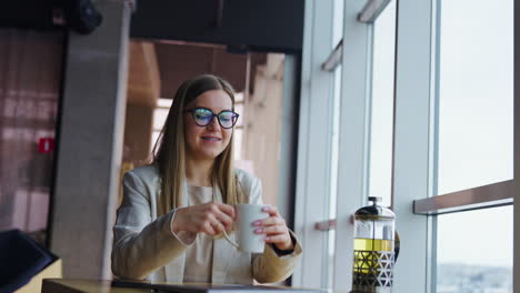 happy girl takes white cup in her hands and smells tea with enjoyment. young woman looks into window and drinks tea.