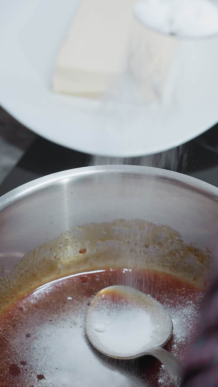 Close-up of cook pouring salt into stew while stirring with a spoon inside pot, various kitchen utensils, including a wooden board and a cookbook, are arranged nearby