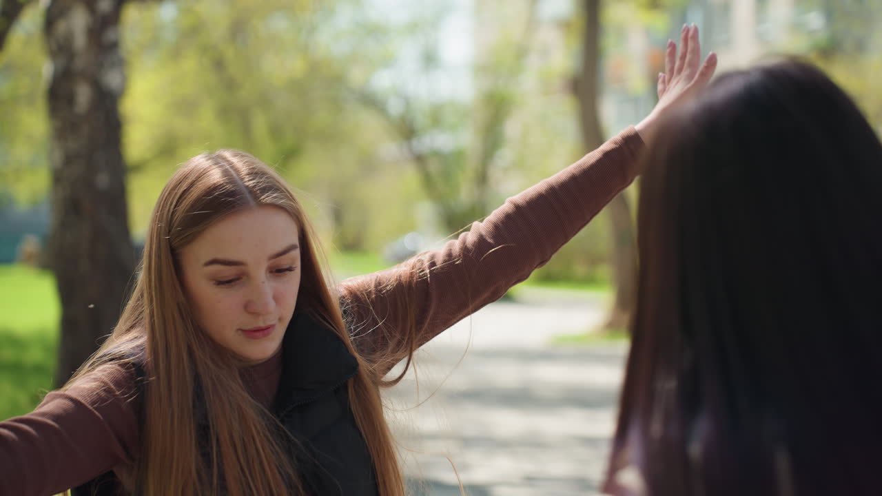Una mujer caucásica levanta los brazos para saludar a una amiga asiática, con energía alegre y una sonrisa brillante, luz solar entre los árboles, ropa de abrigo informal, movimiento expresivo y encuentro espontáneo en un sendero del parque.