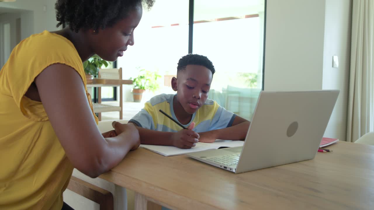 African American mother and son at table leaning, guiding son writing homework with pencil, laptop