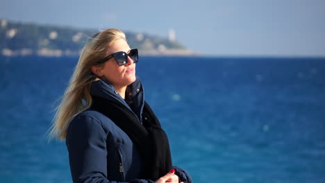 a young woman waves her blond hair in the wind, the blue sea in soft focus behind her on a sunny, windy day slomo