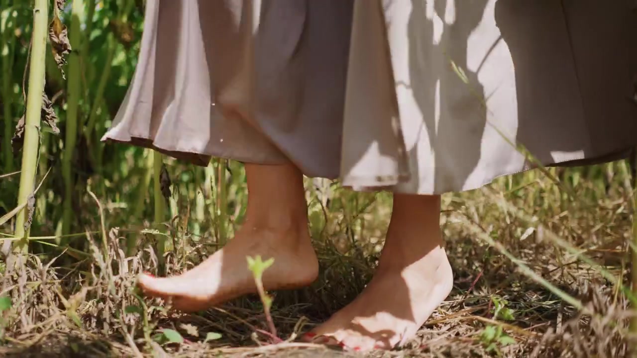 Close up of bare feet walking through tall green plants in natural setting. person wears loose clothing and scene features dry grass beneath their foot with sunlight filtering through leaves
