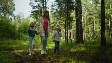 warm moment of mother walking with daughter and son through sunlit forest trail, daughter films surroundings with camcorder while mother points something out to curious boy