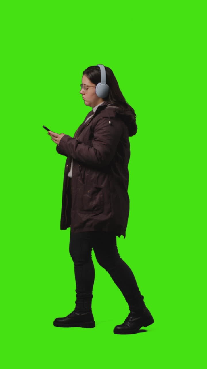 Vertical Video Full Length Shot Of Woman Wearing Wireless Headphones Streaming Music From Mobile Phone Walking Across Frame Against Green Screen