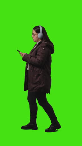 vertical video full length shot of woman wearing wireless headphones streaming music from mobile phone walking across frame against green screen