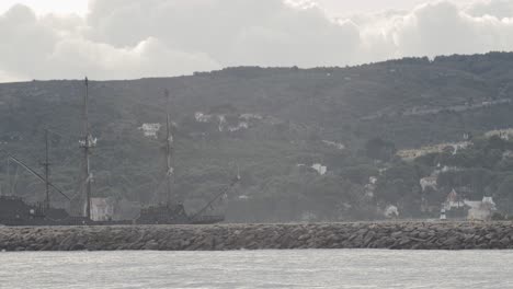 Galeón-Del-Siglo-XVI-Réplica-De-Andalucia-Barco-Que-Llega-Al-Puerto-En-Un-Día-Nublado-Al-Amanecer-Detrás-De-Un-Rompeolas-Con-Montañas-Y-Casas-En-El-Fondo