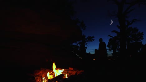 Orange-campfire-burns-with-blue-hour-silhouette-people-under-moon