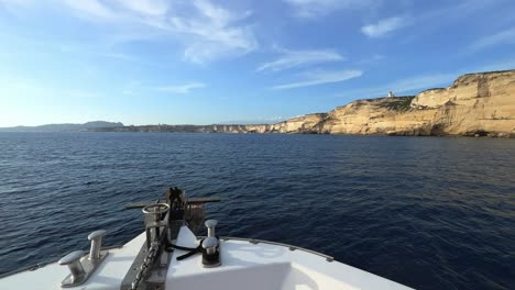 Sailboat-bow-perspective-of-navigating-boat-toward-Corsica-island-cliffs-and-Capo-Pertusato-lighthouse-in-France,-50fps