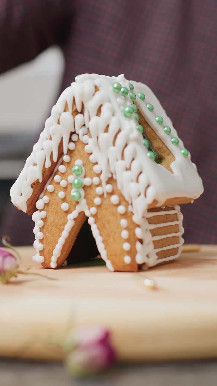panadero girando cuidadosamente y mostrando una casa de pan de jengibre bellamente decorada con hielo blanco y cuentas verdes, bandeja de madera, salpicaduras doradas, flores secas y luz de vela borrosa en el fondo