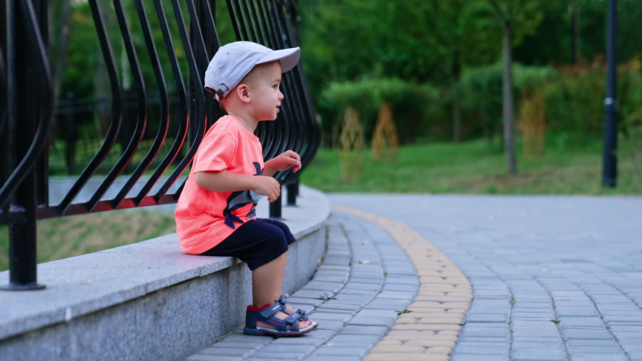 Beautiful baby boy in a cap sits on the edging near the fence. Active kid steps his feet and stands up.