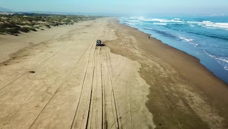 Truck-driving-on-sandy-beach-next-to-ocean-from-Drone