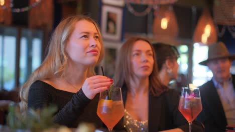 two stylish women are sipping on vibrant cocktails while laughing and chatting at a chic cafe adorned with blooming flowers. the lively atmosphere radiates joy and friendship, making it a perfect summer outing filled with delightful conversations and refreshing drinks