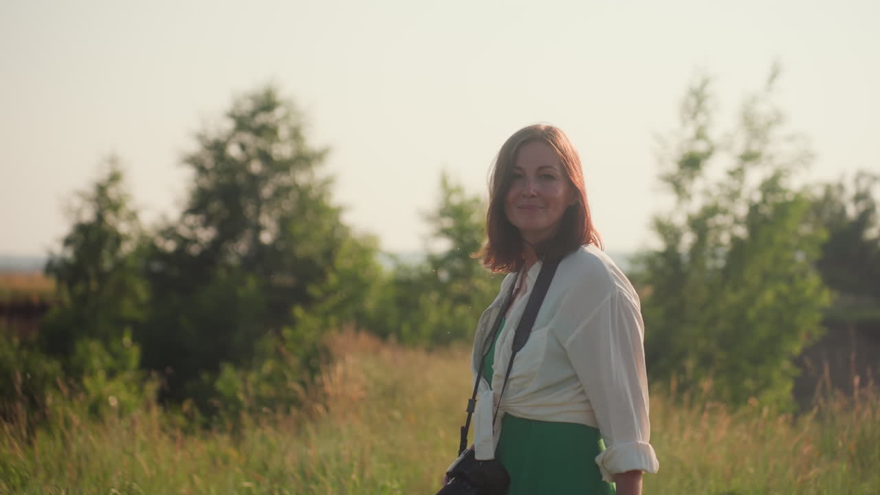 side view of smiling woman wearing camera on neck walking through grassy field under golden sunlight, relaxed expression and casual attire evoke peaceful moment surrounded by summer nature