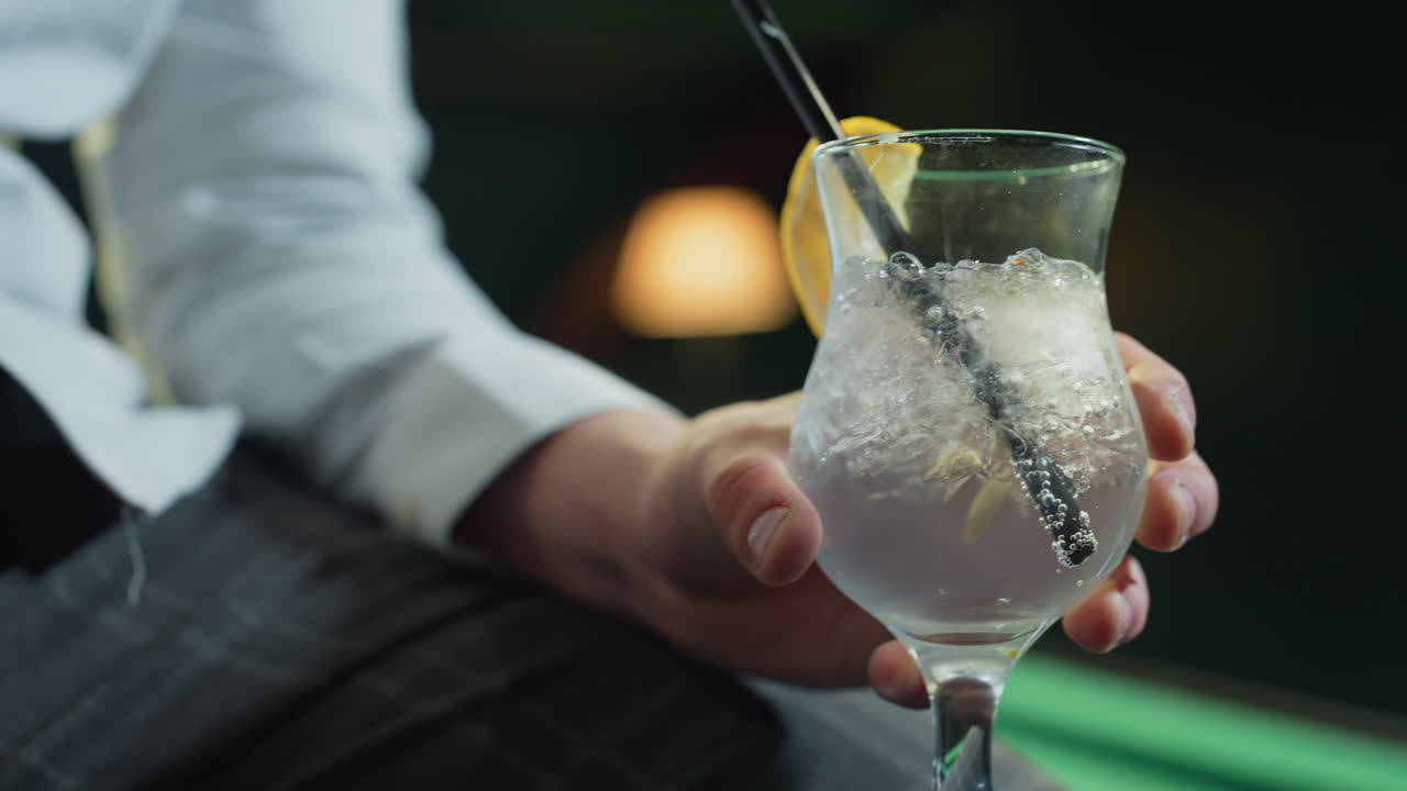 Primo piano di un uomo, volto non visibile, che indossa una camicia bianca e pantaloni grigi e tiene in mano un bicchiere di limonata con una cannuccia nera. Cubetti di ghiaccio e una fetta di limone aggiungono dettagli rinfrescanti, creando un'atmosfera rilassata.