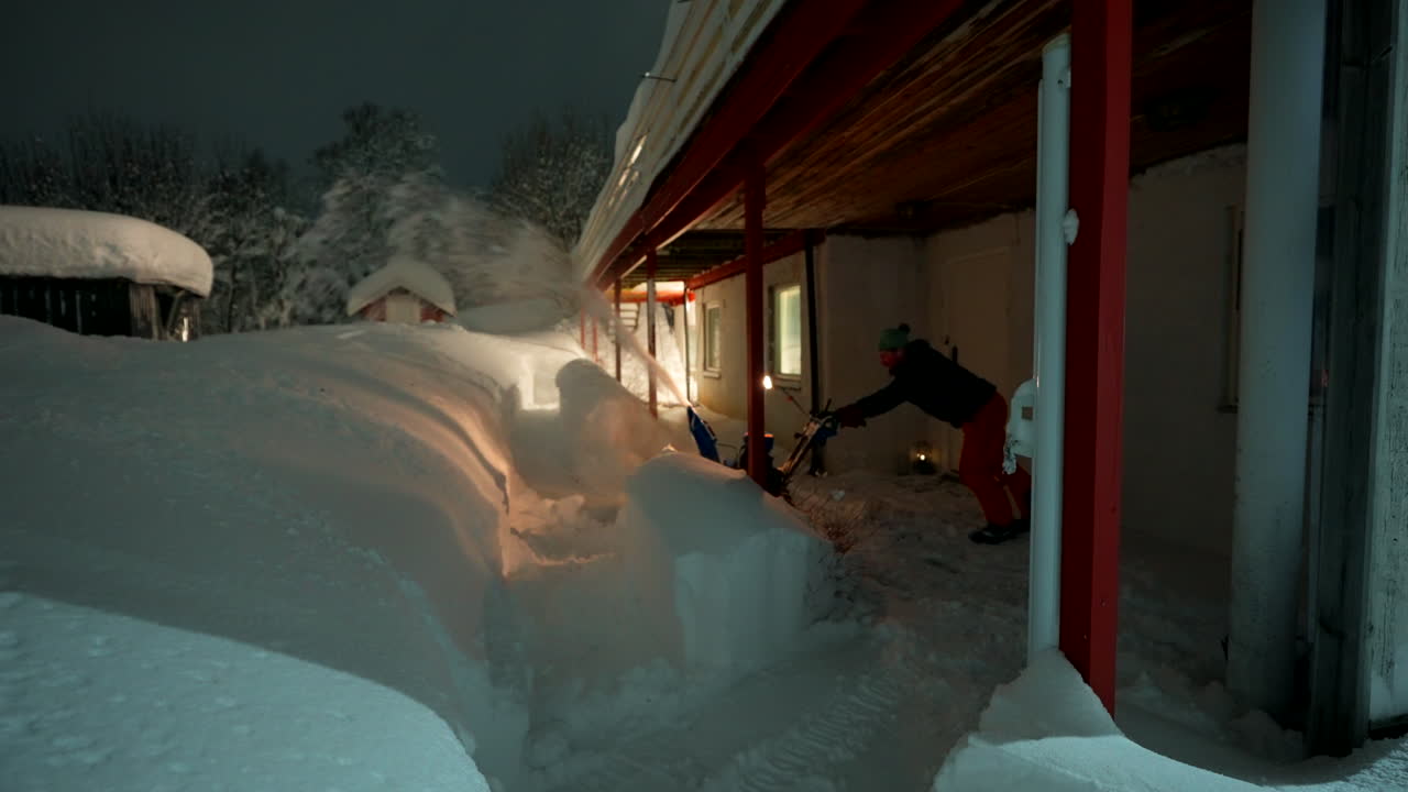 A person walks through heavy snow operating snowplow pushing with great strength to clear path around building at night