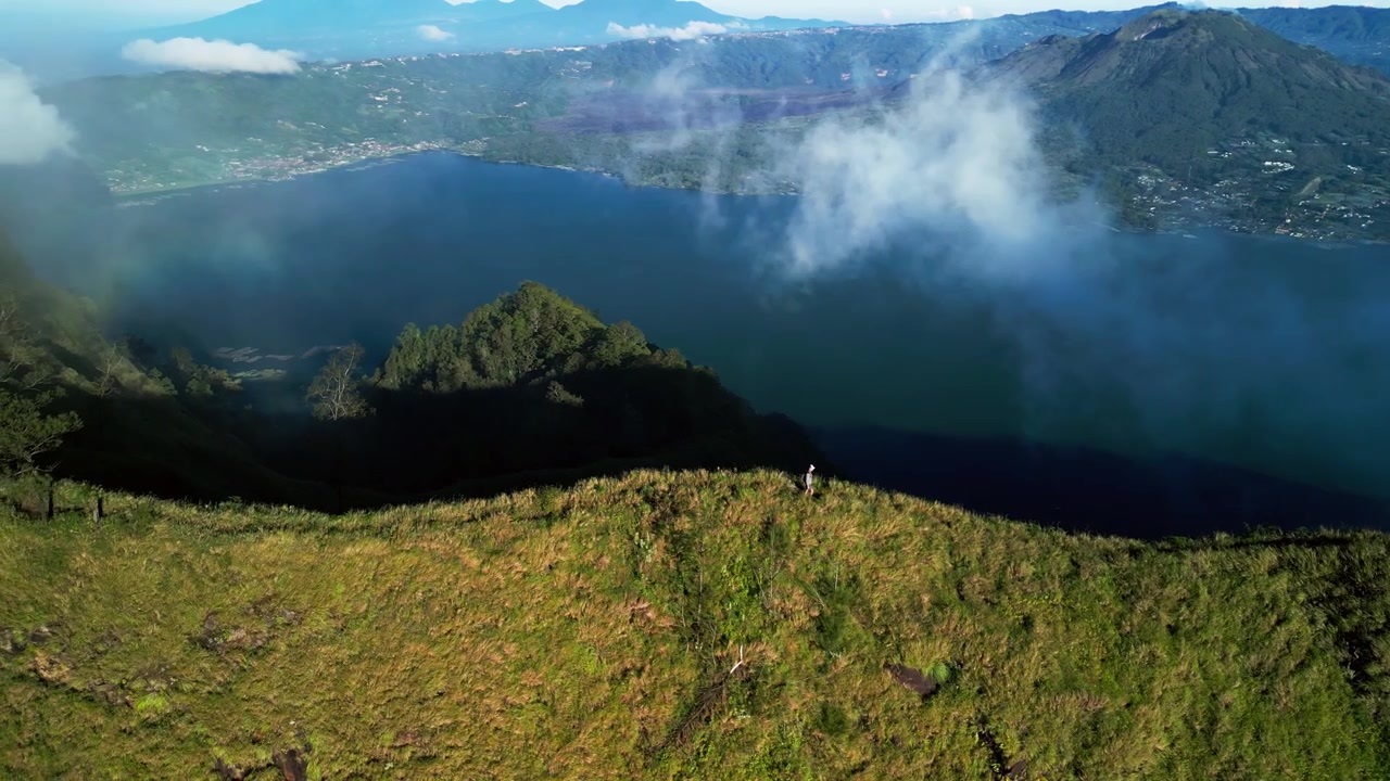 Escursionista solitario che cammina su una cresta esposta sopra il lago del cratere del Monte Batur a Bali, con nuvole che attraversano un paesaggio montano tropicale, riprese aeree fluide con traiettoria ad arco.