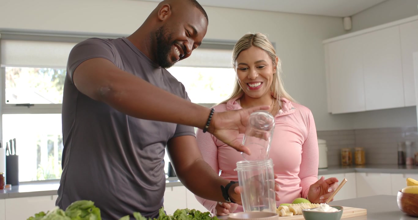 Casal com roupa esportiva na ilha da cozinha, homem despejando copo medidor no liquidificador para nutrição.