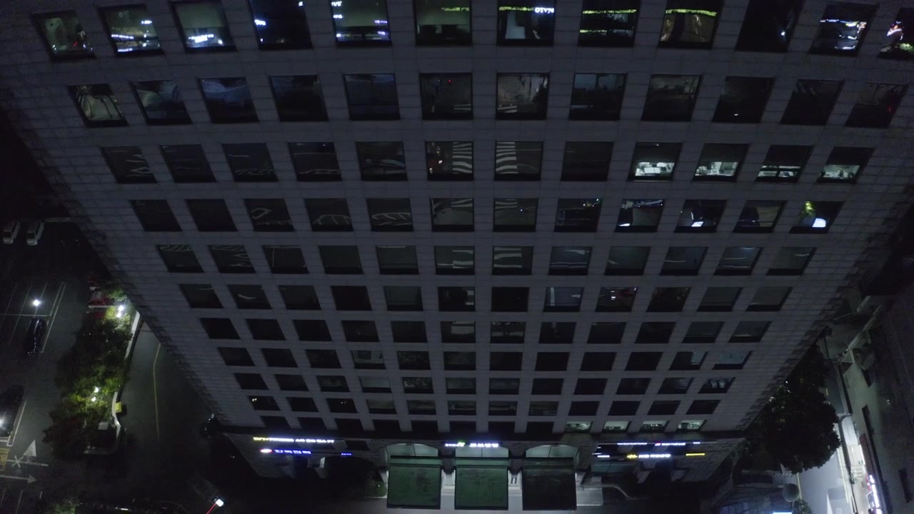 La cámara mira hacia abajo la fachada de un alto edificio de oficinas en la oscuridad, mientras una cuadrícula de ventanas revela espacios de trabajo iluminados dispersos y pisos vacíos