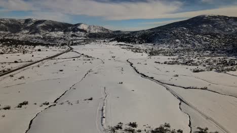 Drohnenflug-Auf-Einer-Schneebedeckten-Ebene,-Bei-Dem-Man-Rillen-Im-Schnee-Sieht,-Die-Durch-Wasserströmungen-Entstanden-Sind,-Und-Auf-Einer-Seite-Befindet-Sich-Eine-Straße-Ohne-Fahrzeuge-Mit-Einem-Hintergrund-Von-Bergen-Im-Winter-In-Avila,-Spanien