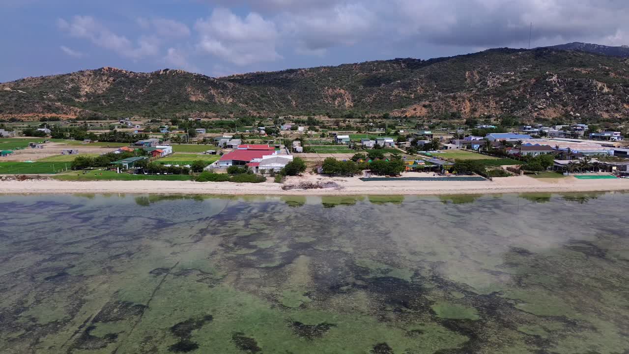 Drone footage flying from the sea towards the beach in Mỹ Hòa, Phan Rang. Right-to-left coastal motion reveals houses, shoreline, and soft waves on a sunny day.