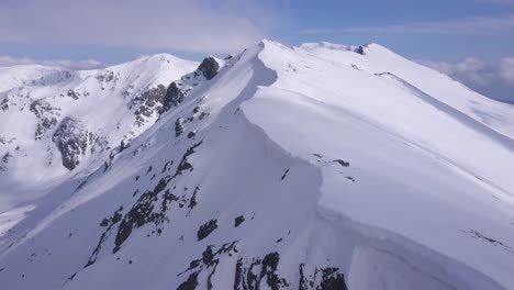 Drohne-Fliegt-Hoch-über-Dem-Schneebedeckten-Bergrücken-Mit-Einem-Gesims-Aus-Schnee,-Das-über-Dem-Felsvorsprung-Hängt