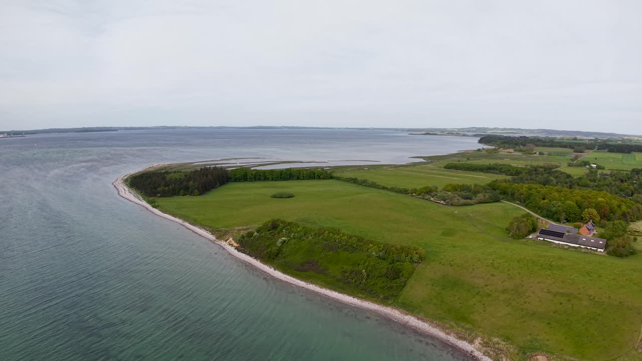 Drone footage of a gently curving coastline with green fields meeting calm sea under a cloudy sky