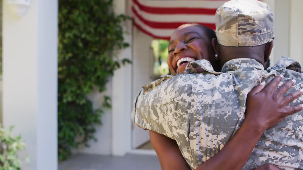 soldado afroamericano abrazando a su esposa sonriente frente a la casa con la bandera estadounidense