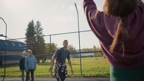 coach slams volleyball across net during training session with two other players watching from behind, lady in maroon sweater playing, outdoor court with distant building in background