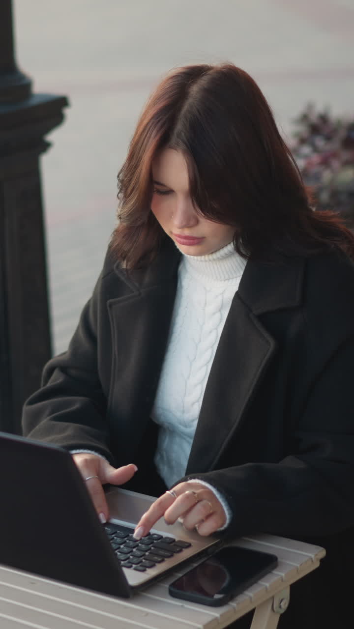 Lady working on laptop outdoors in urban setting, wearing stylish black coat, with decorative black pillar nearby, blurred red brick building in background, and flower gently swaying in breeze