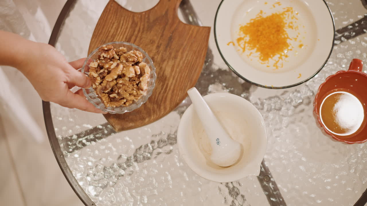 Dall'alto, guarnizione per torta di zucca con noci, mano che posiziona una ciotola di noci pecan tritate su un tagliere di legno, mortaio e scorza d'arancia nelle vicinanze, tavolo di vetro texturizzato, movimento nella calda cucina di casa, azione casuale del fornaio