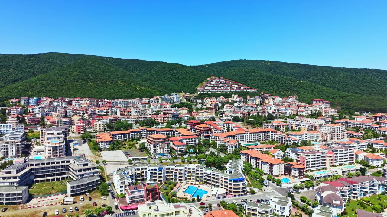 Gündüz vakti, parlak mavi gökyüzü altında, arka planda dağların bulunduğu bir sahil kasabasındaki yerleşim alanı manzarası.